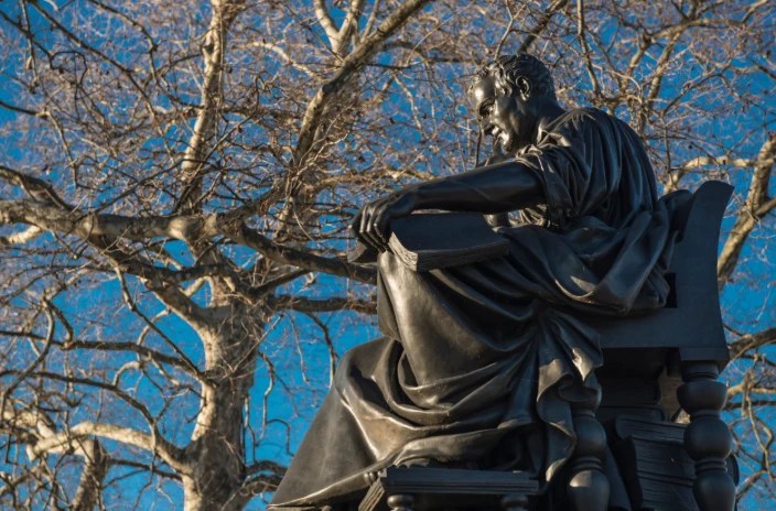 Bronze statue of a seated man holding a book, against bare branches and a blue sky